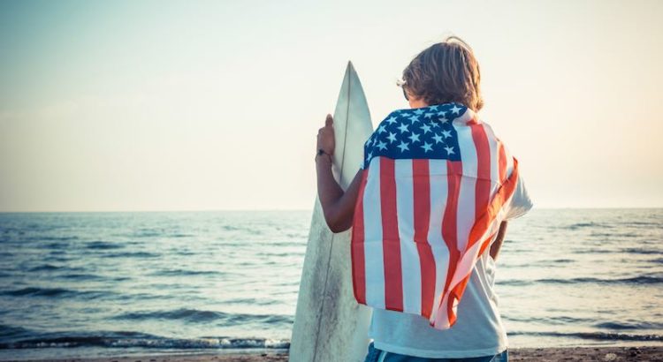 surfer facing the ocean with a surfboard in hand and USA flag over shoulders